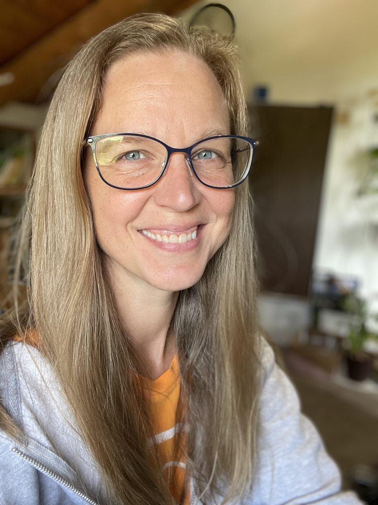 Home 28 Portrait of Esther Yunkin, registered nurse and founder of Health with Hashimoto's, smiling warmly while wearing stylish blue-framed glasses, a gray zip-up hoodie with orange accents, and long blonde hair. She's standing in a cozy home office with wooden beams, green plants, and soft natural light.